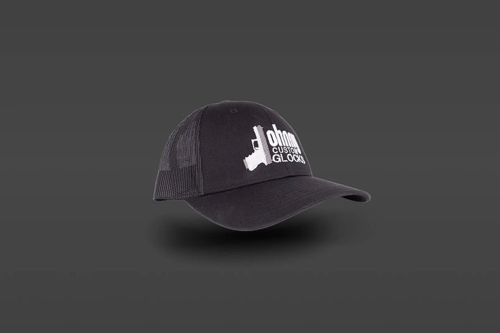A black trucker-style baseball cap with a white embroidered logo on the front, displayed against a dark gray gradient background.